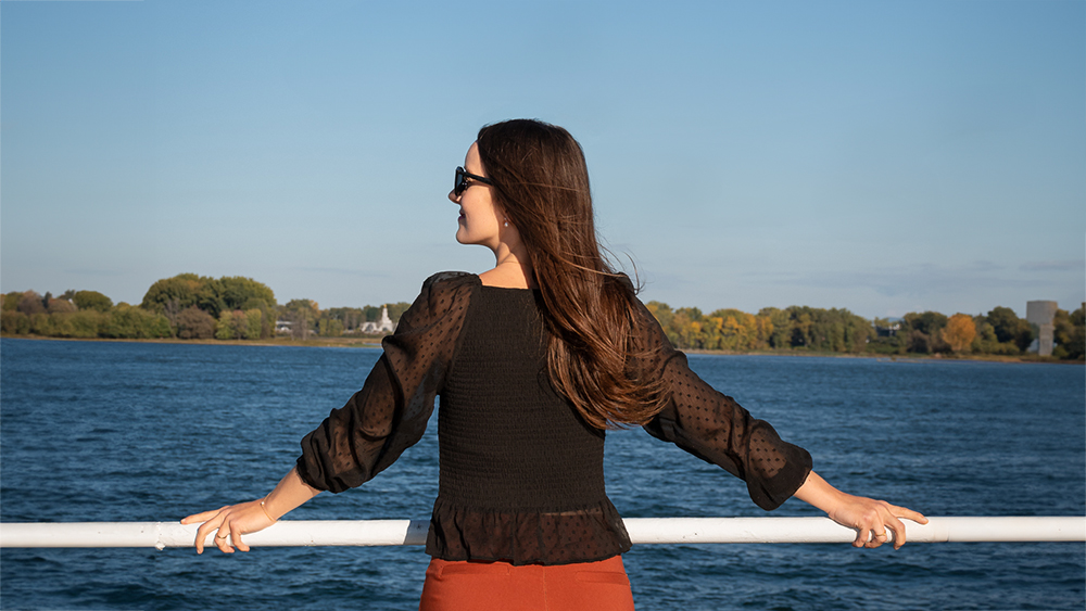 A woman viewed from behind, wearing a black blouse and sunglasses, leaning on a railing, admiring a river with trees and a clear sky in the background.