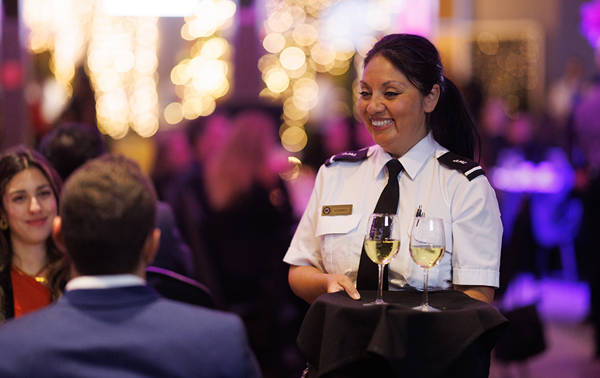 A smiling waitress in uniform serves two glasses of white wine to a table in an elegant and lively setting, illuminated by blurred decorative lights in the background.