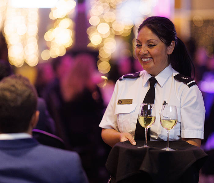 A smiling waitress in uniform serves two glasses of white wine to a table in an elegant and lively setting, illuminated by blurred decorative lights in the background.