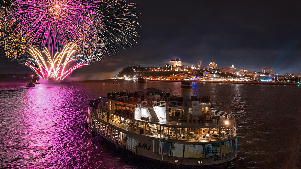 A boat filled with passengers sails on a river under a night sky, as a spectacular fireworks display lights up the sky with pink and golden bursts. In the background, the city is brightly lit, featuring iconic buildings and a castle on a hill.