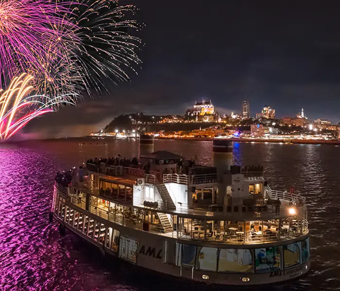 Un bateau rempli de passagers navigue sur une rivière sous un ciel nocturne, tandis qu'un feu d'artifice spectaculaire illumine le ciel avec des éclats roses et dorés. En arrière-plan, la ville est éclairée, avec des bâtiments emblématiques et un château sur une colline.