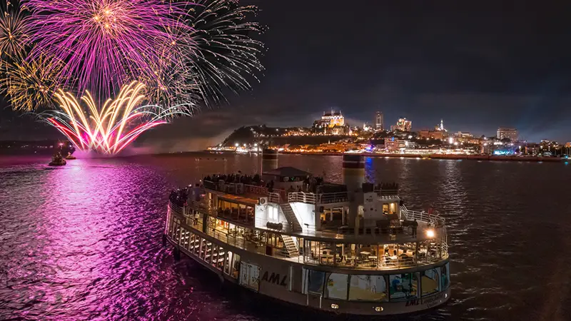 Un bateau rempli de passagers navigue sur une rivière sous un ciel nocturne, tandis qu'un feu d'artifice spectaculaire illumine le ciel avec des éclats roses et dorés. En arrière-plan, la ville est éclairée, avec des bâtiments emblématiques et un château sur une colline.