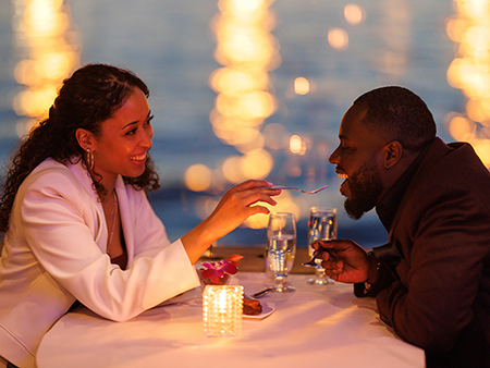 A couple sharing a romantic moment during a candlelit dinner by the water. The smiling woman feeds the man a bite of dessert, with blurred sparkling lights in the background.