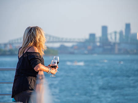  A blonde woman, viewed from behind, holding a glass of red wine, looking at a river with a bridge and skyscrapers in the background on a sunny day.