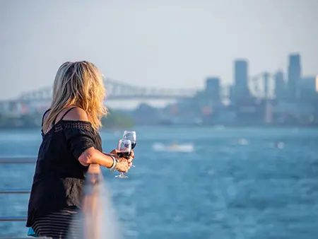 Une femme blonde, de dos, tenant un verre de vin rouge, regardant une rivière avec un pont et des gratte-ciel en arrière-plan par une journée ensoleillée.