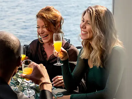 A group of friends enjoying a cheerful moment in a bright restaurant, with a smiling woman holding a glass of mimosa.