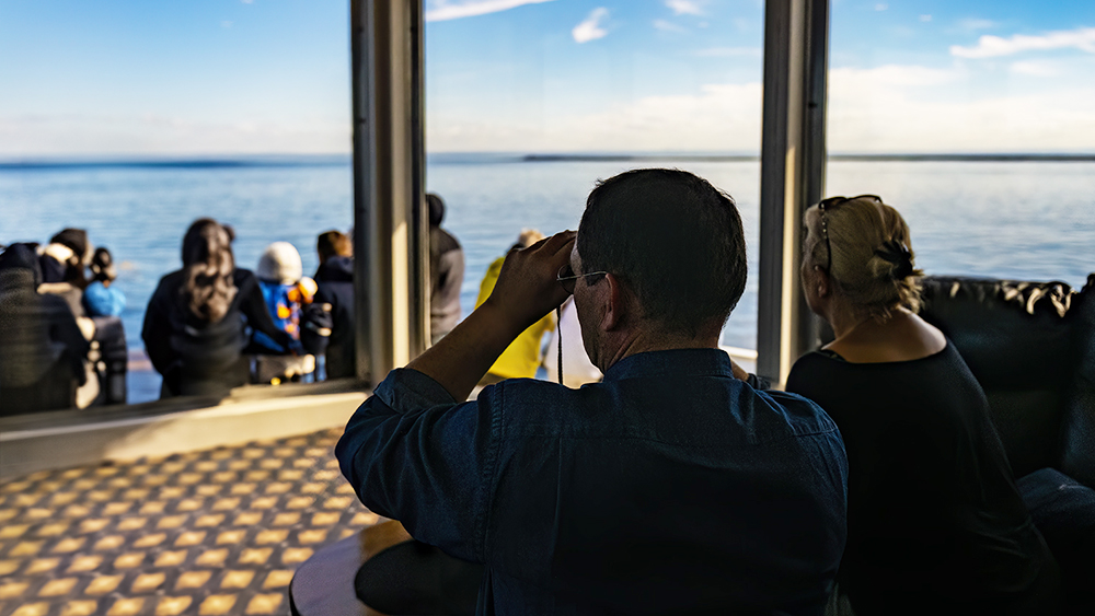 A man observes the horizon through binoculars from inside a glass-paneled cabin, accompanied by a woman sitting beside him. Outside, a group of people watches the calm sea under a blue sky. Sunlight streams through the windows.