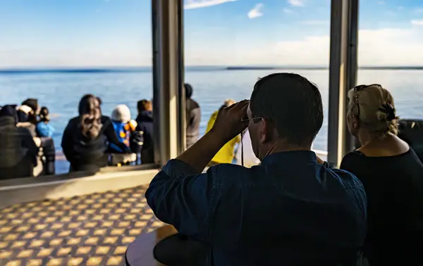 A man observes the horizon through binoculars from inside a glass-paneled cabin, accompanied by a woman sitting beside him. Outside, a group of people watches the calm sea under a blue sky. Sunlight streams through the windows.