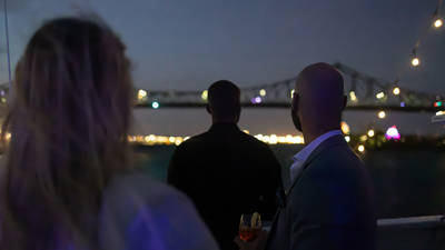 Three people viewed from behind, enjoying an evening on a boat with a lit-up bridge and city lights in the background.