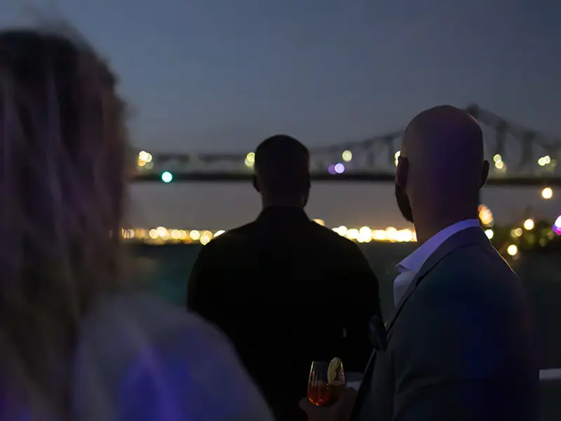 Three people viewed from behind, enjoying an evening on a boat with a lit-up bridge and city lights in the background.