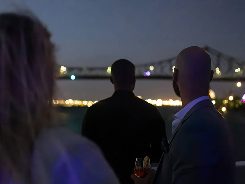 Three people viewed from behind, enjoying an evening on a boat with a lit-up bridge and city lights in the background.