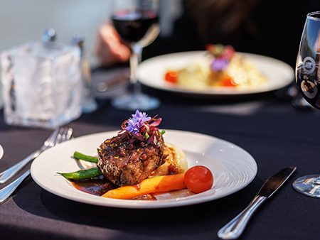 A gourmet steak dish garnished with a flower, paired with a glass of red wine, overlooking the serene backdrop of a water view.