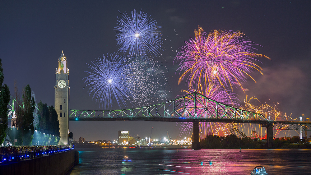 Un feu d'artifice coloré éclate dans le ciel nocturne près du pont Jacques-Cartier et de la tour de l'Horloge, avec des reflets sur le fleuve et une foule observant depuis le quai.