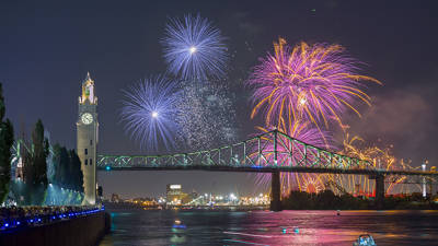 Colorful fireworks burst in the night sky near the Jacques Cartier Bridge and the Clock Tower, with reflections on the river and a crowd watching from the quay.
