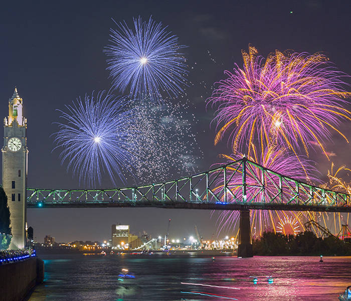Colorful fireworks burst in the night sky near the Jacques Cartier Bridge and the Clock Tower, with reflections on the river and a crowd watching from the quay.