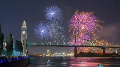 Un feu d'artifice coloré éclate dans le ciel nocturne près du pont Jacques-Cartier et de la tour de l'Horloge, avec des reflets sur le fleuve et une foule observant depuis le quai.