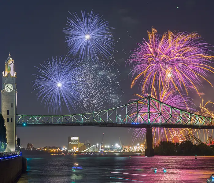 Un feu d'artifice coloré éclate dans le ciel nocturne près du pont Jacques-Cartier et de la tour de l'Horloge, avec des reflets sur le fleuve et une foule observant depuis le quai.