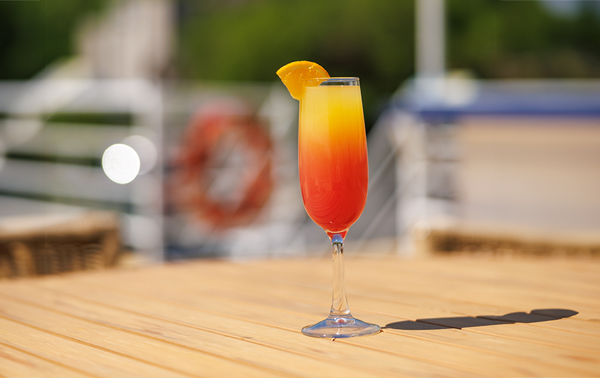 An orange mimosa cocktail in a flute with an orange wedge on top, served on a wooden table on one of the outdoor terraces of the AML Cavalier Maxim ship in Montreal.