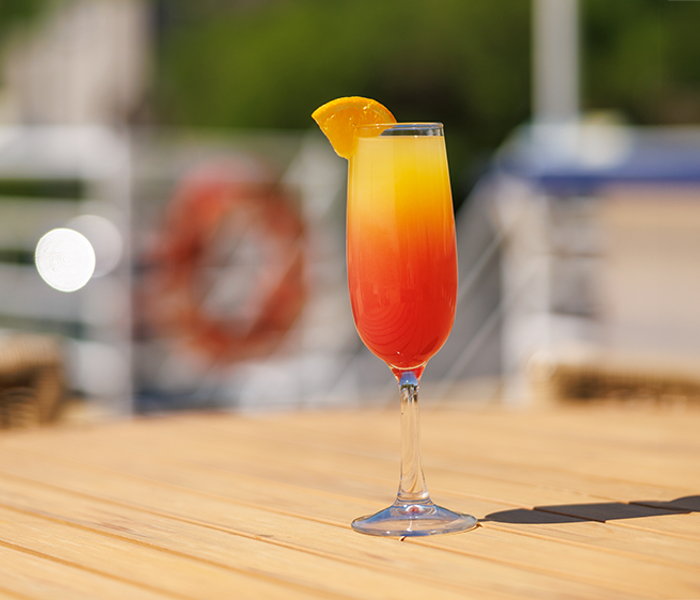 An orange mimosa cocktail in a flute with an orange wedge on top, served on a wooden table on one of the outdoor terraces of the AML Cavalier Maxim ship in Montreal.
