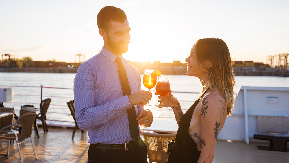 Un couple élégamment habillé trinque avec des cocktails sur le pont d'un bateau au coucher du soleil, avec une rivière et un quai en arrière-plan.