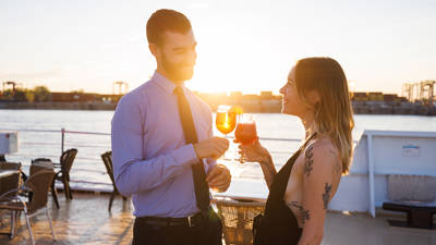 An elegantly dressed couple toasts with cocktails on a boat deck at sunset, with a river and a dock in the background.