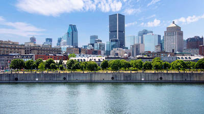 A panoramic view of a city's waterfront with a green park, historic buildings, and modern skyscrapers under a partly cloudy blue sky.