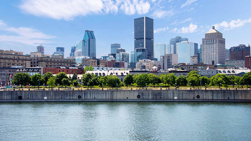 A panoramic view of a city's waterfront with a green park, historic buildings, and modern skyscrapers under a partly cloudy blue sky.