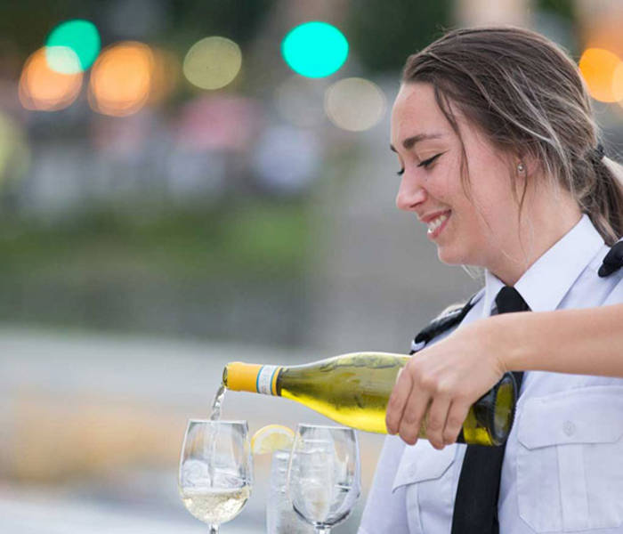 A smiling waitress in uniform pours white wine into glasses, with a blurred background of lights and greenery.