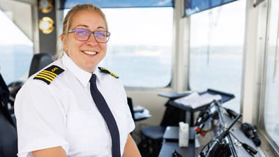 A smiling boat captain, wearing a white uniform with black and gold epaulettes, stands in the ship's cockpit. Through the windows in the background, there is a clear view of the water and sky.