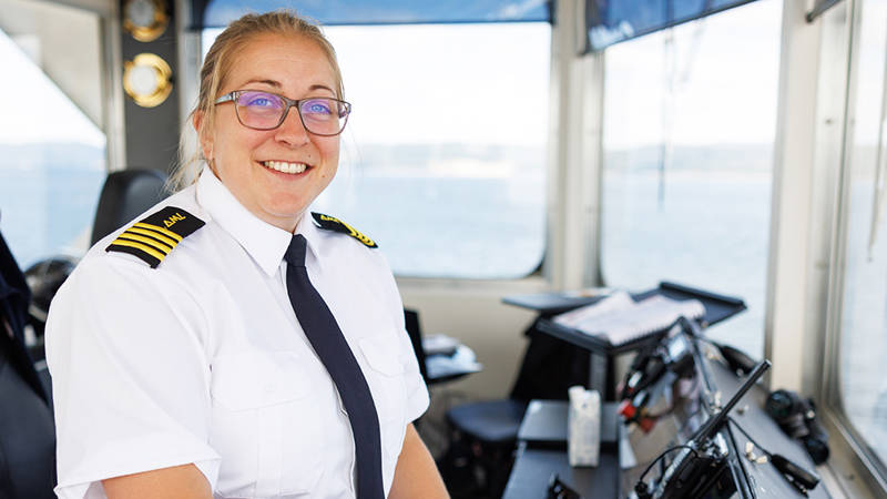 A smiling boat captain, wearing a white uniform with black and gold epaulettes, stands in the ship's cockpit. Through the windows in the background, there is a clear view of the water and sky.