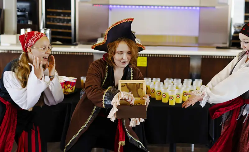 Trois personnes déguisées en pirates interagissent de manière ludique à l'intérieur. L'une d'elles tient un coffre en bois avec une expression de surprise, tandis que les deux autres réagissent avec enthousiasme. Ils se trouvent près d'une table avec plusieurs bouteilles, suggérant un rassemblement ou un événement amusant.