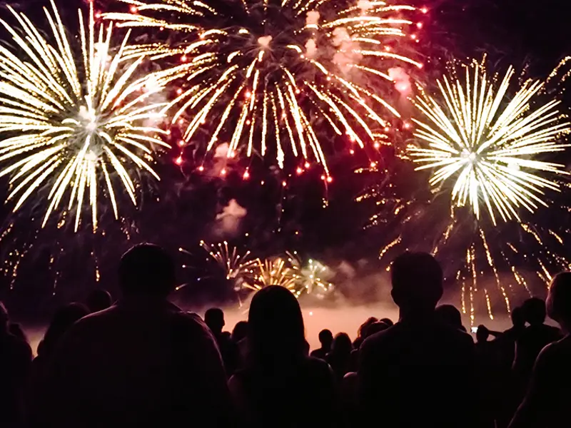 Une foule admire un spectaculaire feu d'artifice illuminant le ciel nocturne avec des éclats dorés et rouges, créant une ambiance festive et magique.