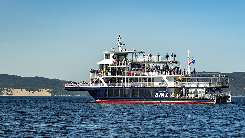 A large cruise ship filled with passengers across multiple decks sails on calm waters. In the background, hills and sand dunes are visible under a clear blue sky.