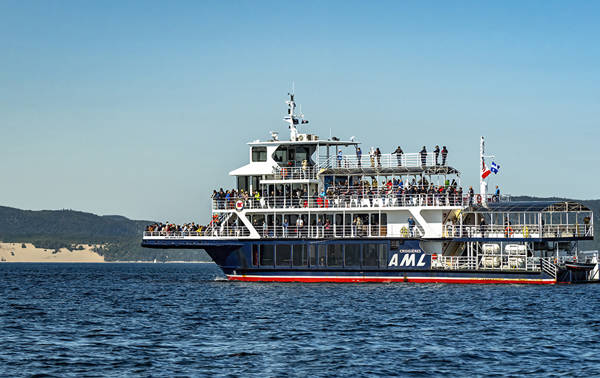 A large cruise ship filled with passengers across multiple decks sails on calm waters. In the background, hills and sand dunes are visible under a clear blue sky.