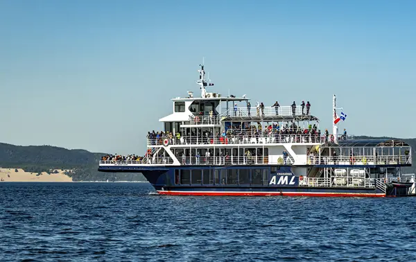 A large cruise ship filled with passengers across multiple decks sails on calm waters. In the background, hills and sand dunes are visible under a clear blue sky.