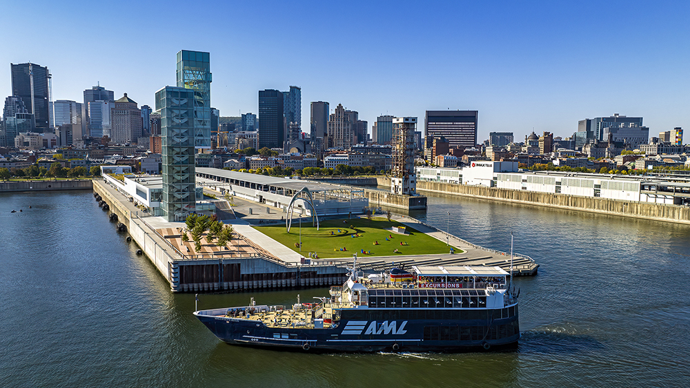 Aerial view of a cruise ship sailing near a modern dock with green spaces and sculptures, surrounded by urban buildings and a river under a clear sky.
