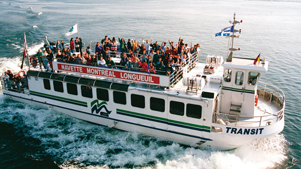 Une navette fluviale pleine de passagers profite d'une traversée entre Montréal et Longueuil sur des eaux calmes, avec des drapeaux flottants sur le bateau.
