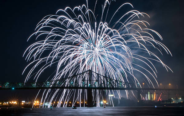 A spectacular fireworks display lights up the night sky above the Jacques Cartier Bridge, with its reflections shimmering on the river.
