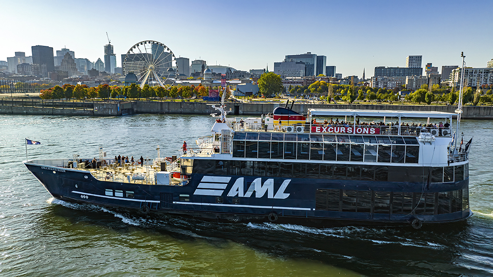 Un grand bateau de croisière noir avec l'inscription "AML" naviguant sur une rivière. En arrière-plan, une grande roue, des arbres d'automne et des bâtiments urbains sous un ciel bleu dégagé.
