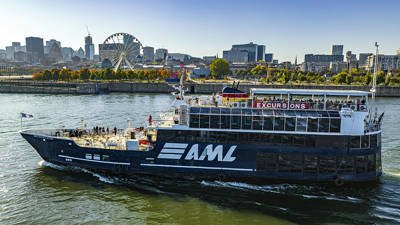 A large black cruise ship with "AML" written on it, sailing on a river. In the background, a Ferris wheel, autumn trees, and urban buildings under a clear blue sky.