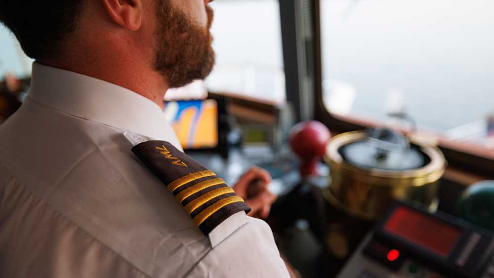 Gros plan sur un capitaine en uniforme avec des galons dorés sur l'épaule, dans la cabine de pilotage d'un bateau, entouré d'équipements de navigation et d'instruments.