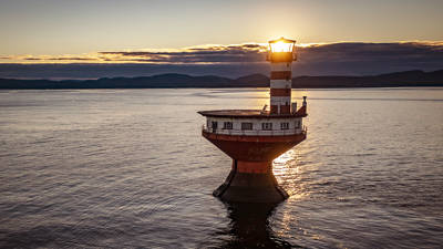 A lone, striped lighthouse stands in calm waters during sunset. The sun shines brightly behind the lantern, casting a warm glow. Dark, distant hills and a partly cloudy sky serve as a backdrop.