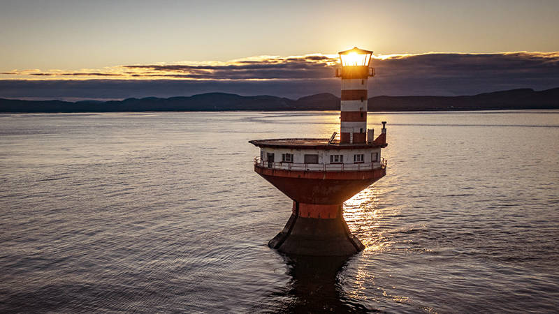 A lone, striped lighthouse stands in calm waters during sunset. The sun shines brightly behind the lantern, casting a warm glow. Dark, distant hills and a partly cloudy sky serve as a backdrop.