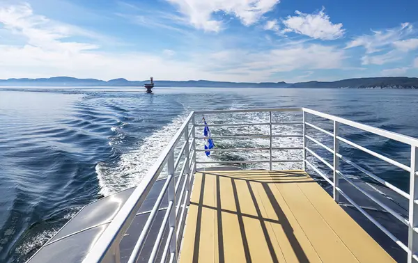 View from the deck of a boat sailing on a calm blue sea. The wake of the boat trails behind, and a buoy is visible in the water. The horizon shows distant hills under a bright blue sky with scattered clouds.
