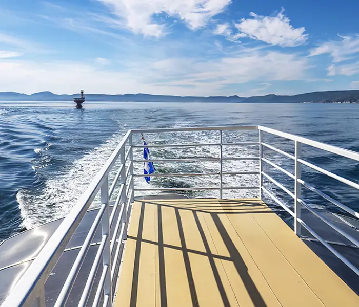 Vue depuis le pont d'un bateau naviguant sur une mer bleue et calme. Le sillage du bateau s'étire derrière lui, et une bouée est visible dans l'eau. À l'horizon, on aperçoit des collines lointaines sous un ciel bleu éclatant parsemé de quelques nuages.