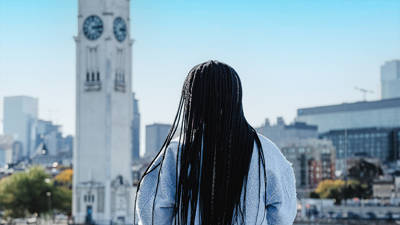  A woman viewed from behind with long black braids, looking at a tall white clock tower and a cityscape with modern buildings under a clear blue sky.