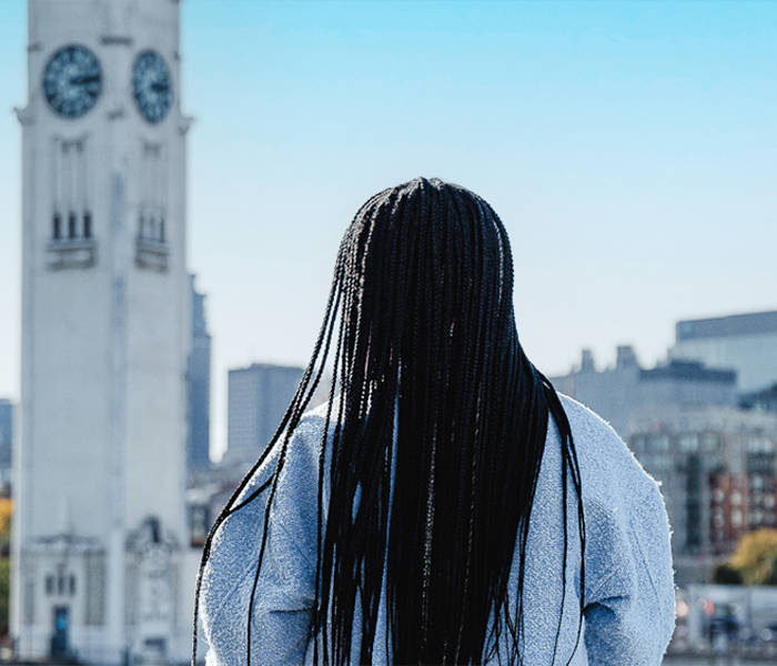  A woman viewed from behind with long black braids, looking at a tall white clock tower and a cityscape with modern buildings under a clear blue sky.