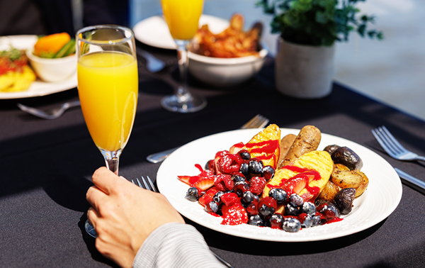 Gros plan sur une assiette brunch végane avec une saucisse végane, des fruits et des pommes de terre grelots avec une main tenant un verre de mimosa.