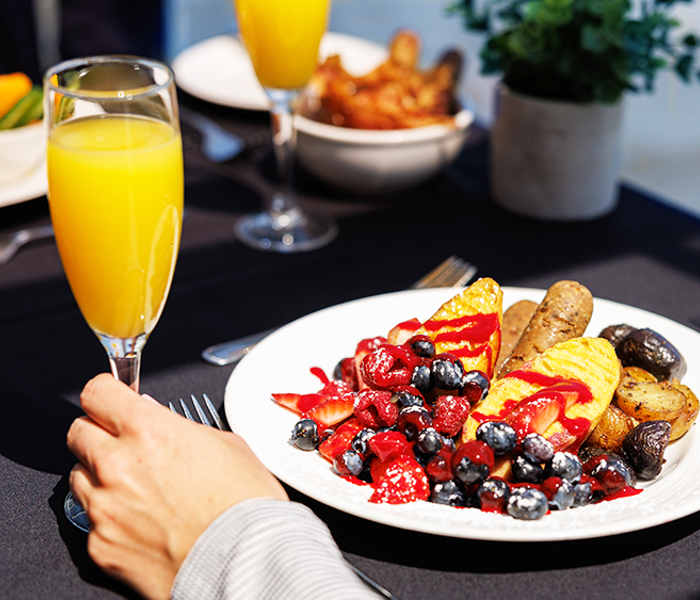 Gros plan sur une assiette brunch végane avec une saucisse végane, des fruits et des pommes de terre grelots avec une main tenant un verre de mimosa.