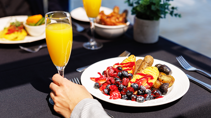  A close-up of a vegan brunch plate with vegan sausage, fruits, and baby potatoes, with a hand holding a mimosa glass.